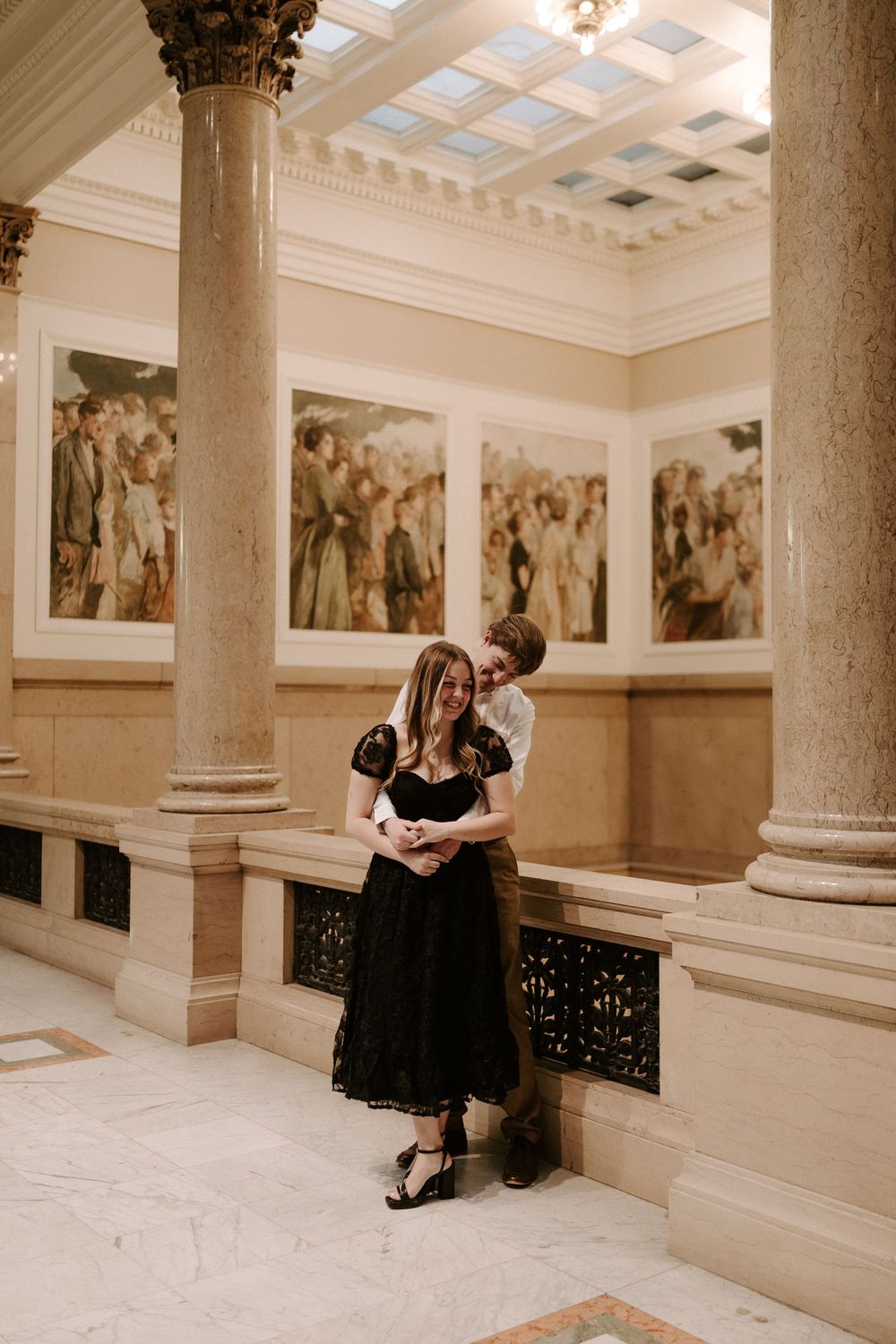 Man embracing woman from behind near marble columns with classical paintings in elegant building.