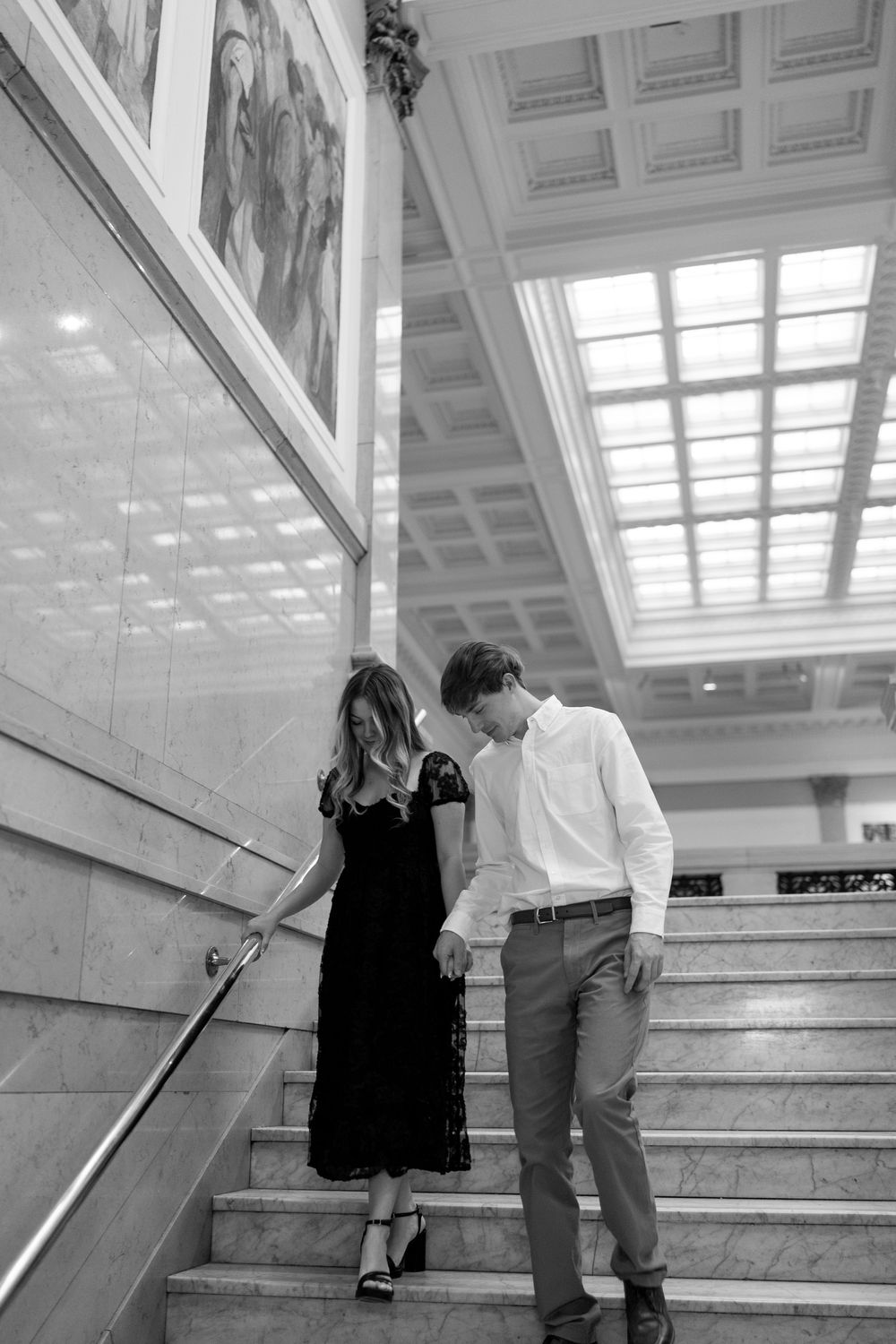 Black and white photo of couple walking down marble stairs in grand building with murals.