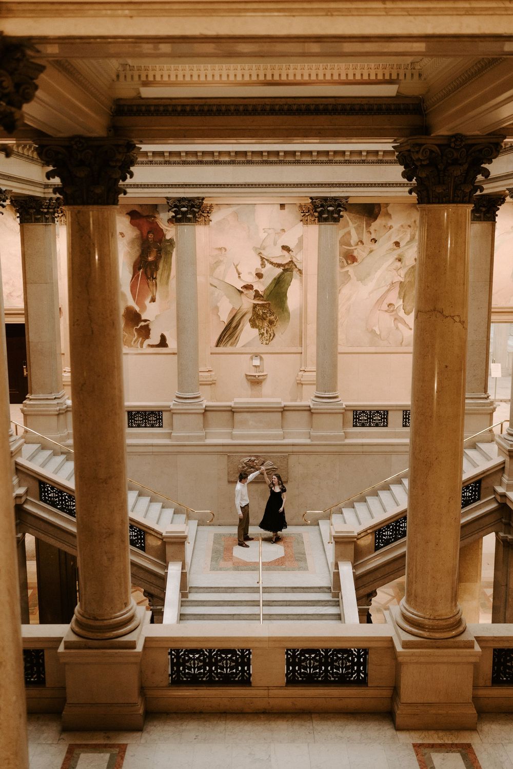 Couple stands on grand marble staircase in ornate building with classical murals and columns overhead.
