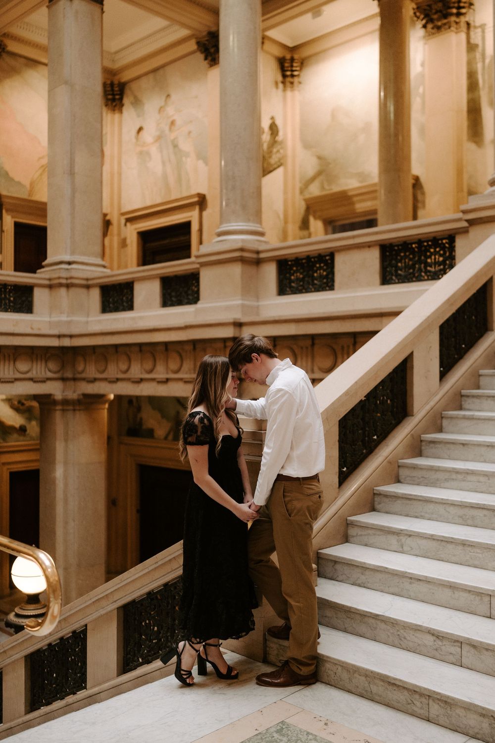 Couple shares romantic kiss on elegant marble staircase with classical columns and murals behind them.