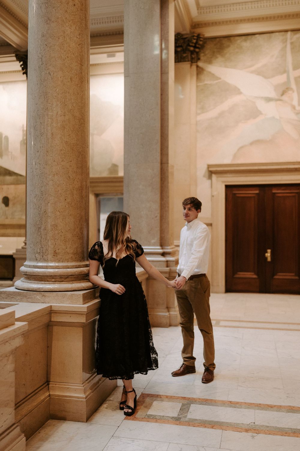 Woman in black lace dress leans against marble column as man in white shirt holds her hand.