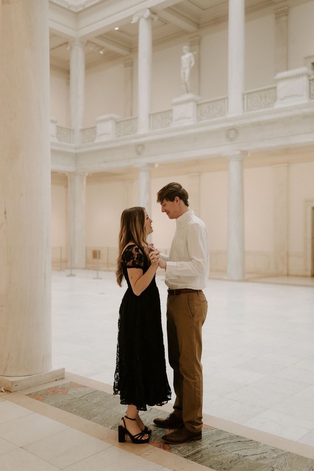 Couple holds hands and gazes at each other in grand white marble hall with tall columns.