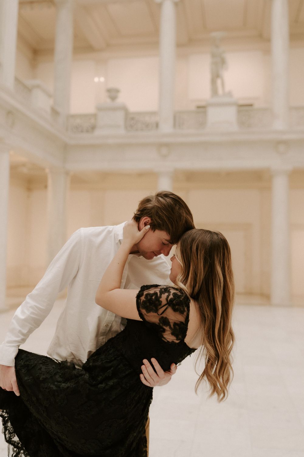 Couple shares romantic dip and kiss in elegant white marble hall with classical architecture.