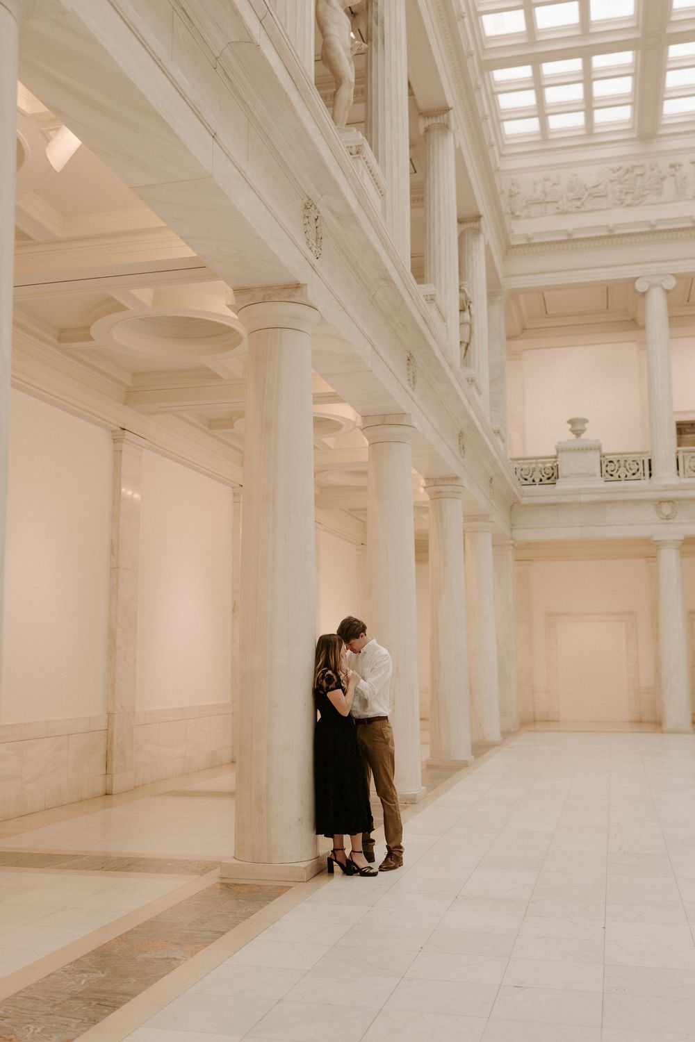 Couple shares tender moment leaning against tall white marble column in grand classical hallway.
