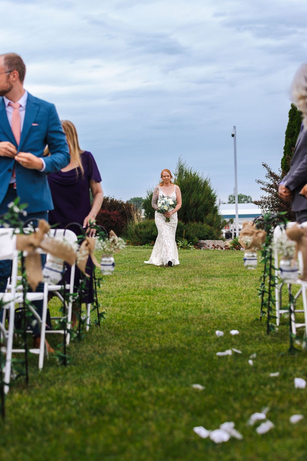 Guests seated in white chairs on a grassy lawn for an outdoor wedding ceremony with scattered flower petals.