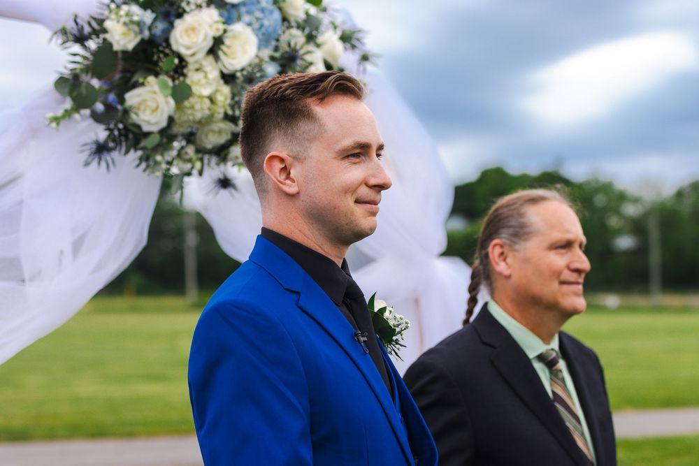 Two people in formal attire stand outdoors with blue sky and green grass in the background.