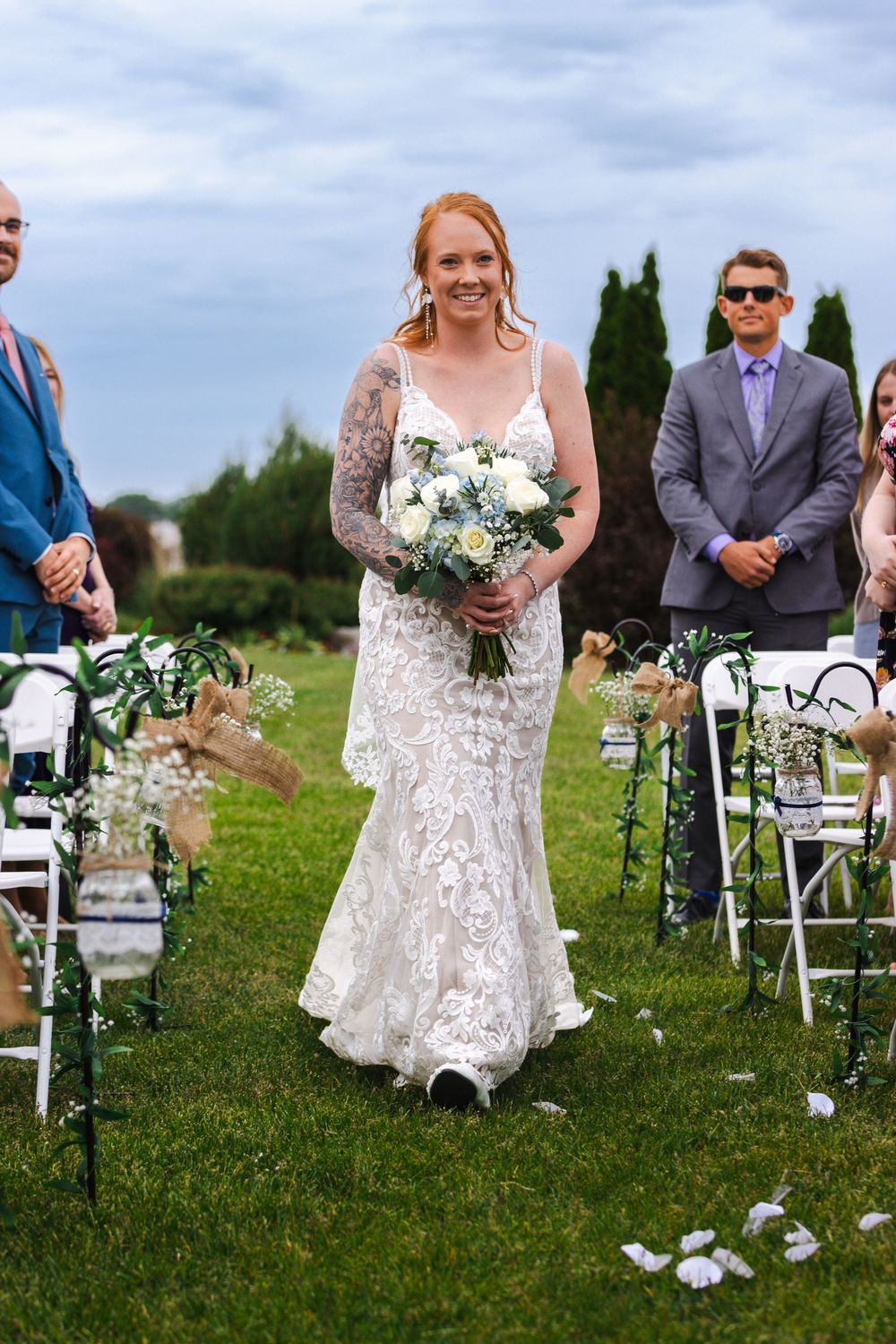 A bride in a white lace dress walks down an outdoor aisle lined with white chairs on a grassy field.