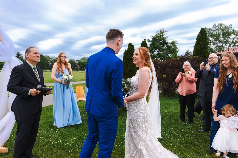 A wedding celebration outdoors with guests gathered on a grassy lawn under blue skies during a summer afternoon.