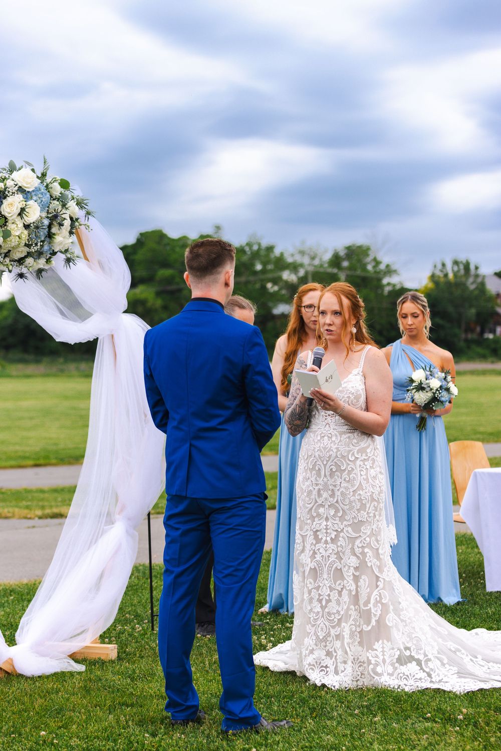 A wedding party stands outdoors with bridesmaids in light blue dresses and a bride in a lace gown with flowing veil.