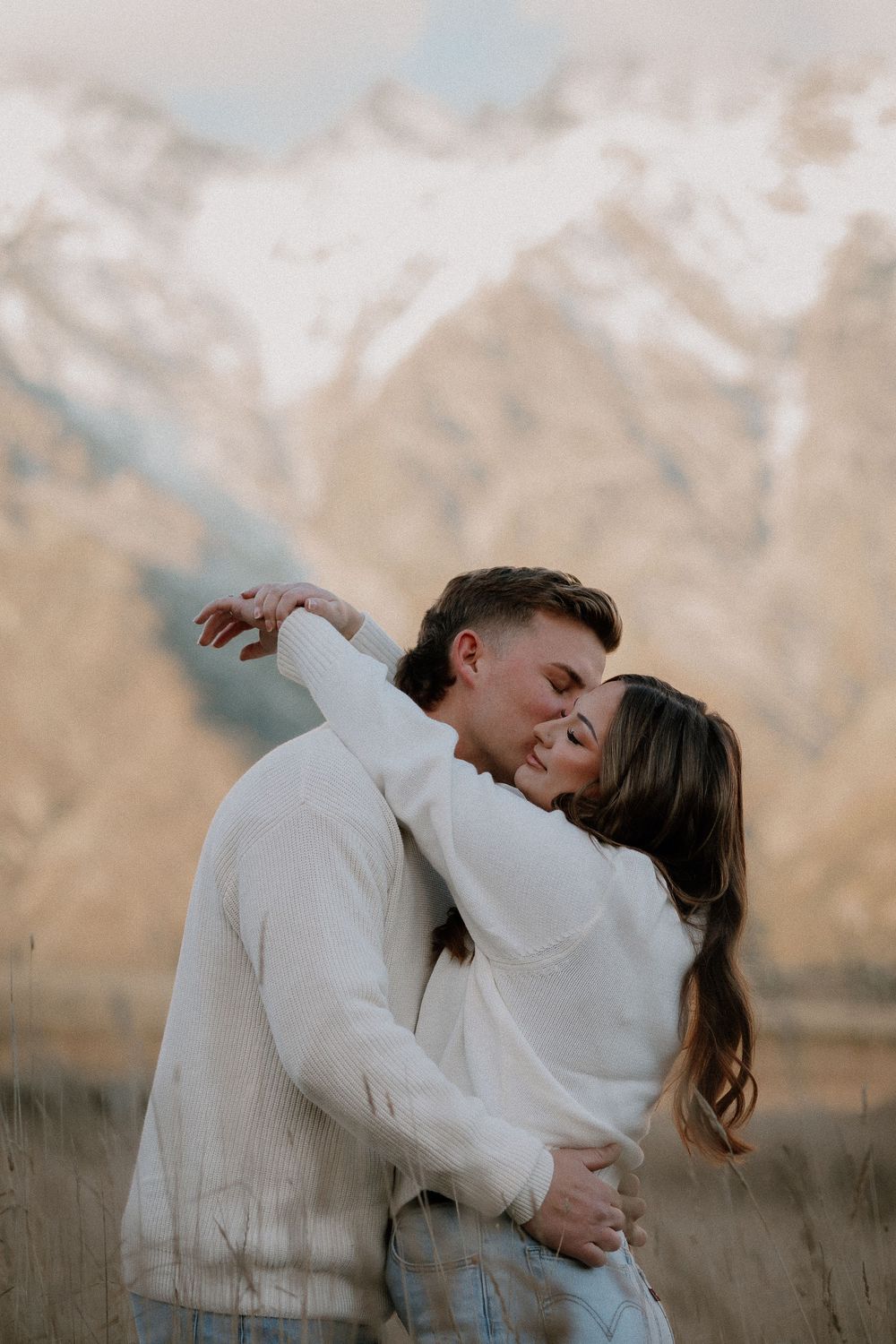 Couple embracing and smiling in front of snow-capped mountains, wearing white sweaters in a romantic outdoor engagement photo.