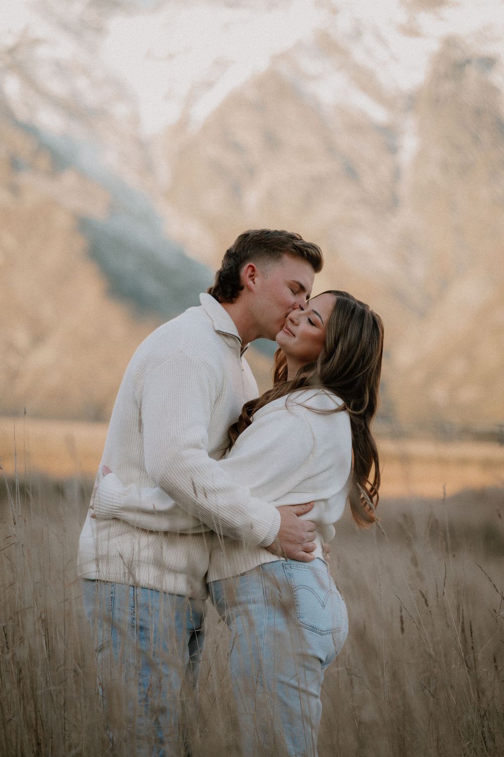 Couple embracing in golden field with snow-capped mountains, wearing cream sweaters and jeans, man kissing woman's cheek.
