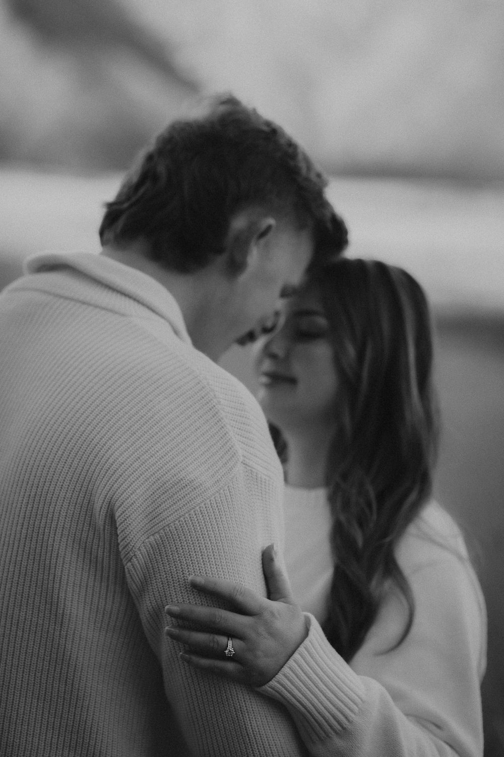 Black and white photo of a couple sharing a tender embrace, woman wearing an engagement ring resting her hand on mans chest.