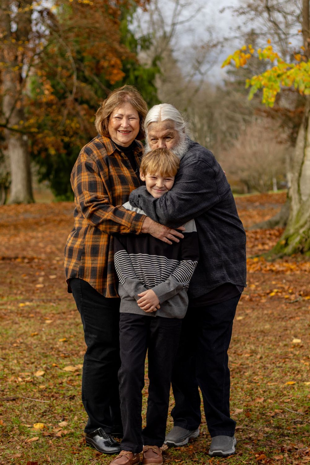 Grandparents hugging young grandson in colorful autumn park setting, smiling warmly together.