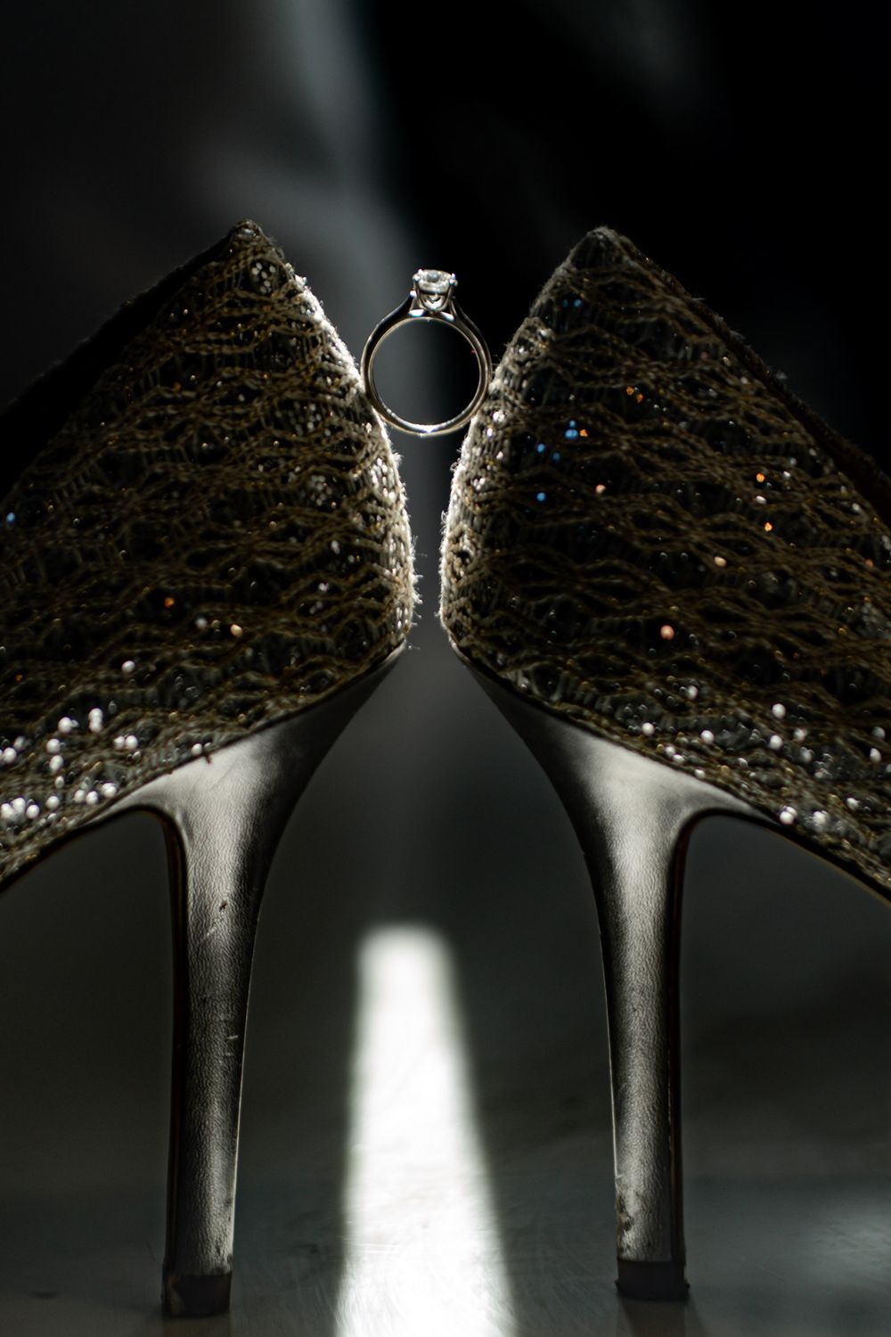 Diamond engagement ring balanced between two glittering gold high heel wedding shoes in dramatic lighting.