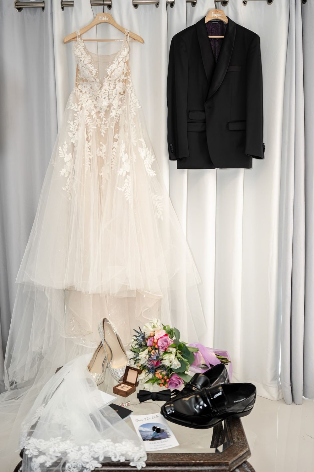 Wedding flat lay featuring lace bridal gown, black tuxedo, bouquet, heels, and dress shoes on display.
