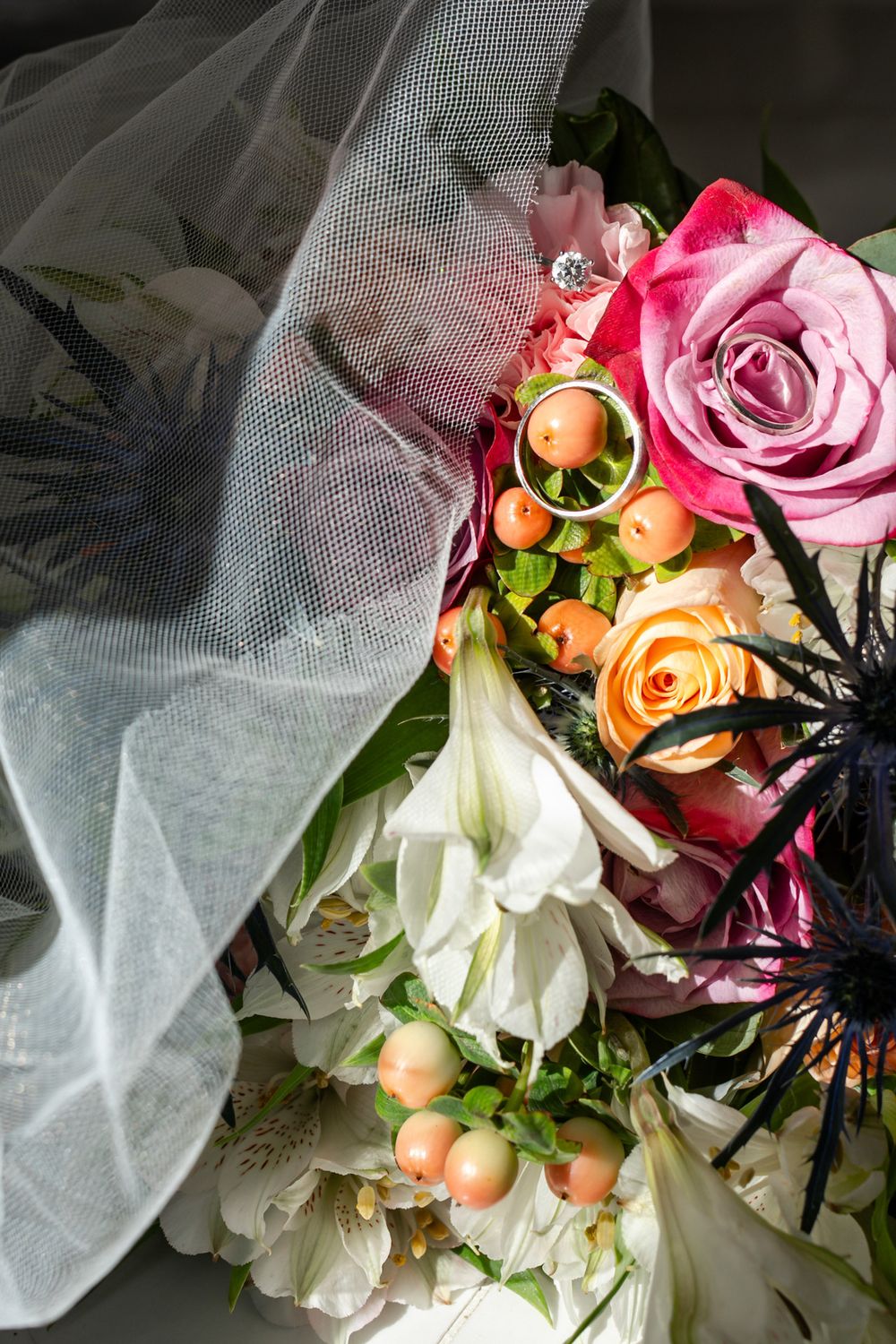 Colorful bridal bouquet with pink and orange roses, white flowers, and wedding rings resting on veil.