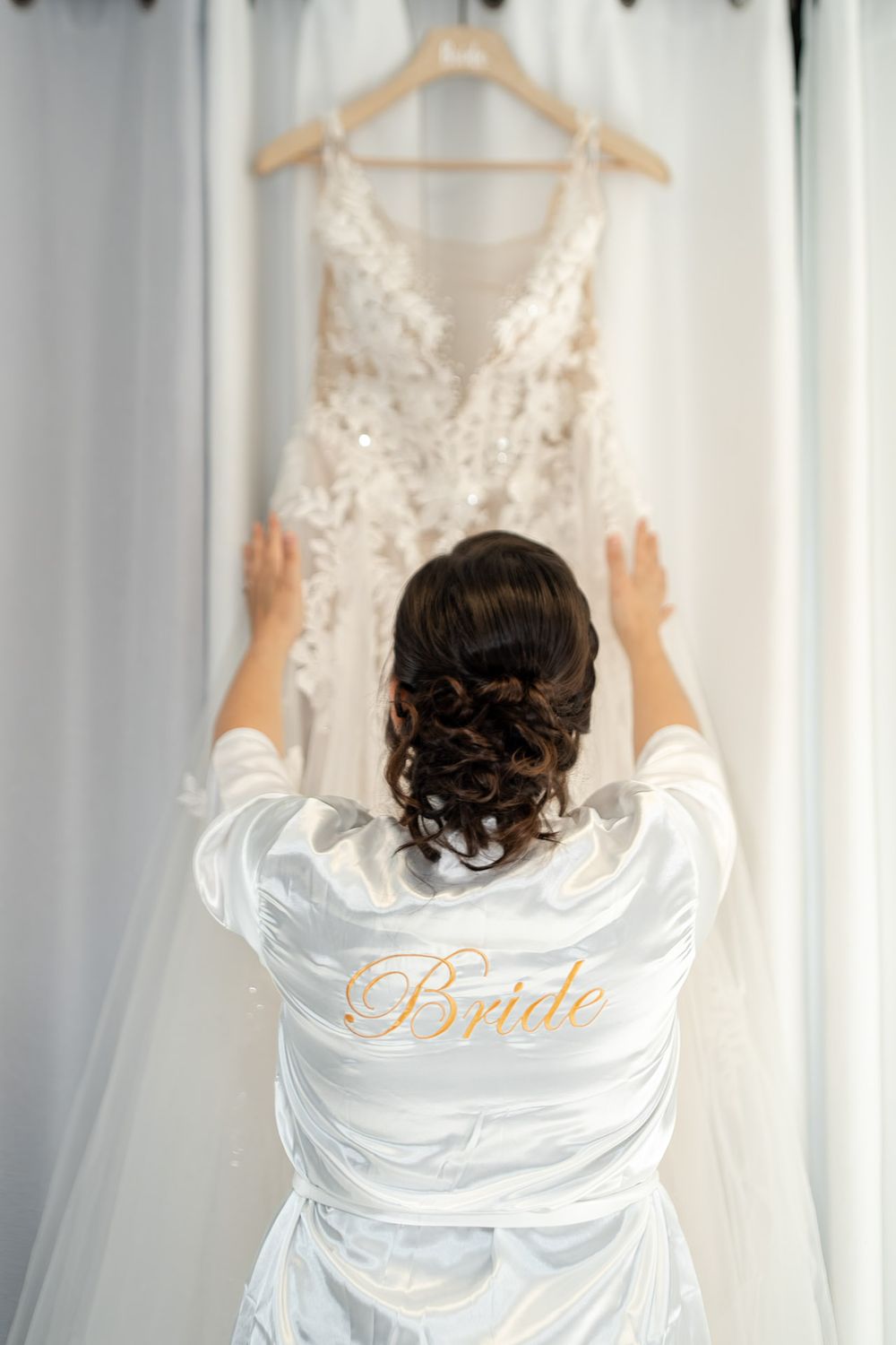 Bride in white satin robe reaching for her lace wedding gown hanging on a curtain rod.