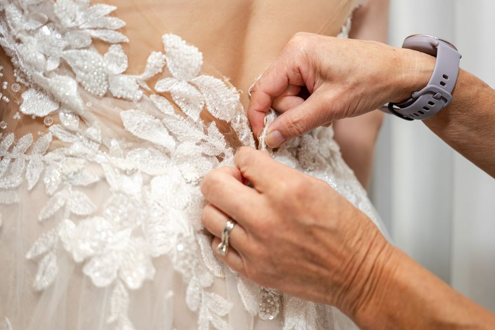 Close-up of hands carefully fastening delicate buttons on the back of an intricate lace wedding gown.