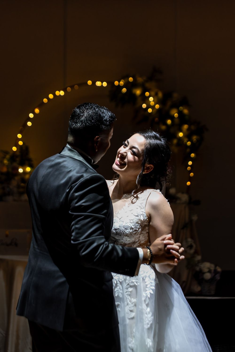 Bride in lace gown laughs joyfully with groom during first dance before bokeh light arch.