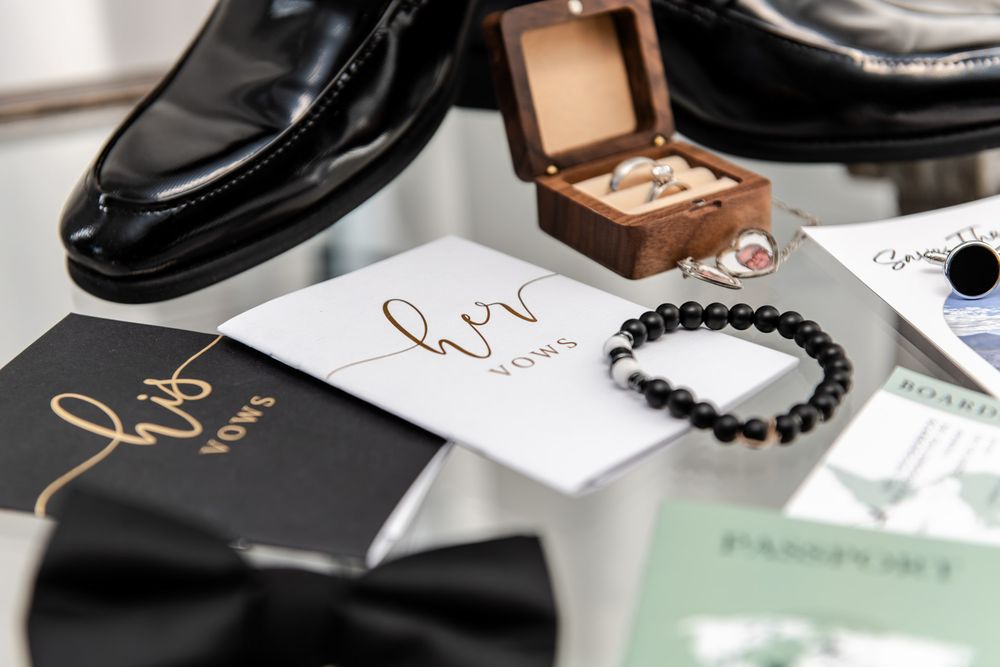 Wedding flat lay featuring his and her vow books, bow tie, rings, passport, and dress shoes.
