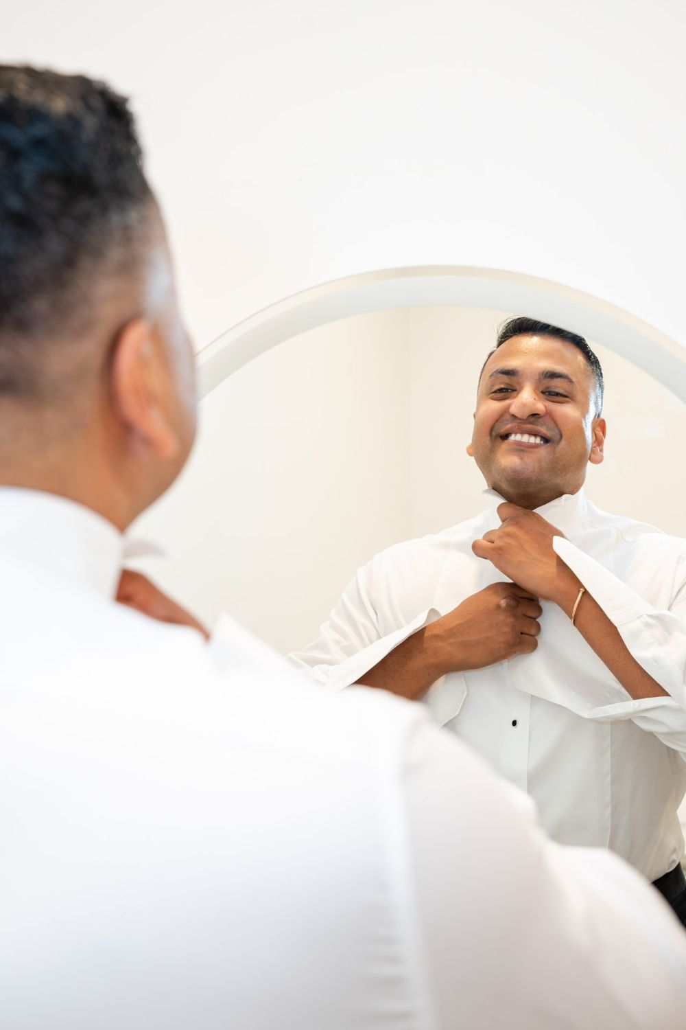 Smiling groom adjusts collar in oval mirror while getting ready on wedding day.