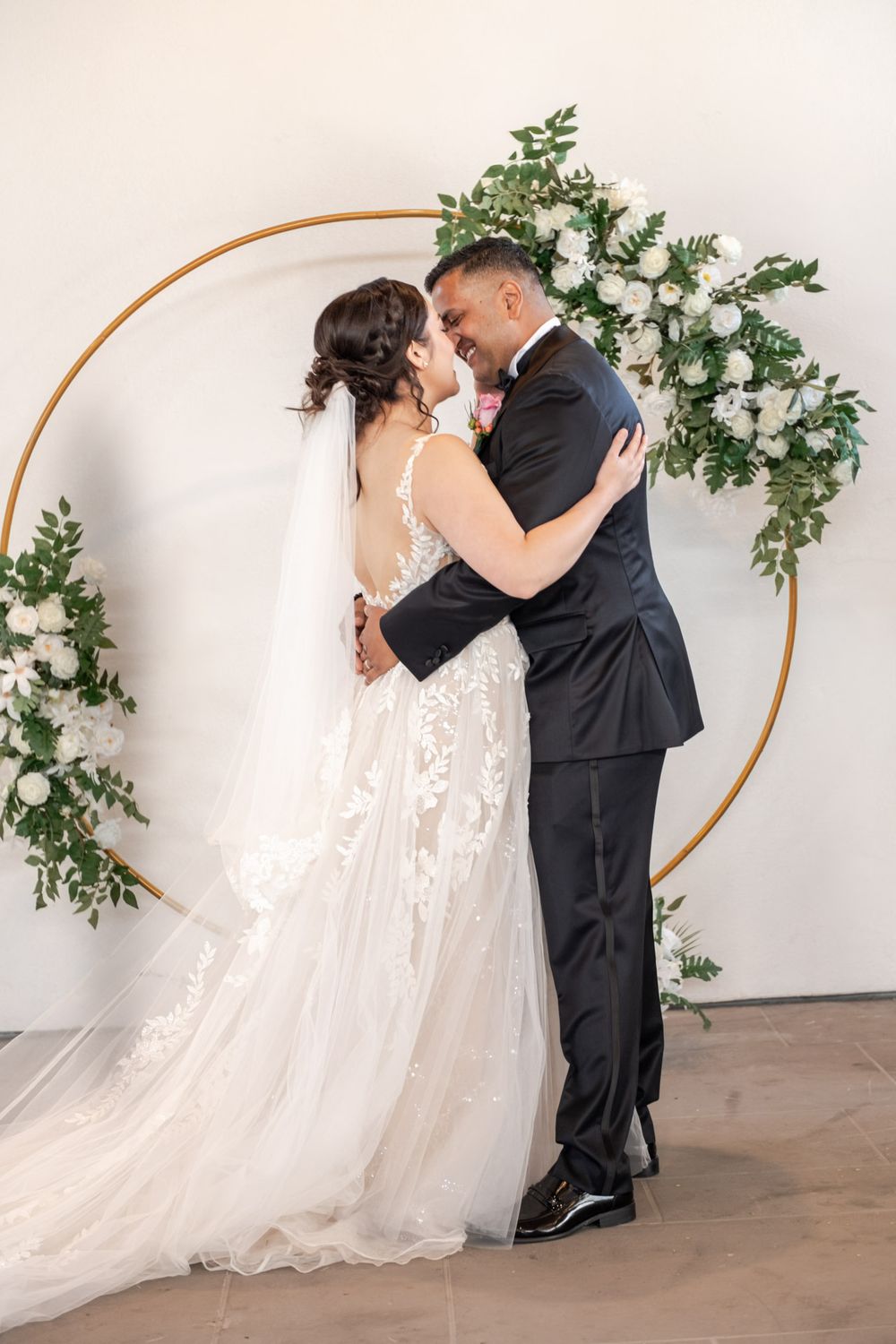 Bride and groom embrace and smile before a gold circular arch adorned with white roses and greenery.