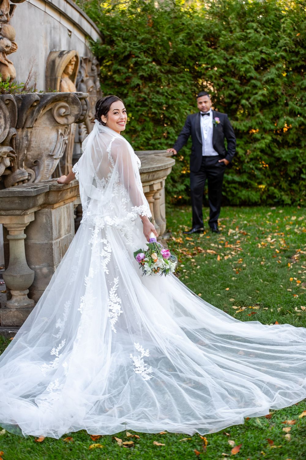 Bride in stunning lace gown with long cathedral veil and train smiling over shoulder, groom standing in background.