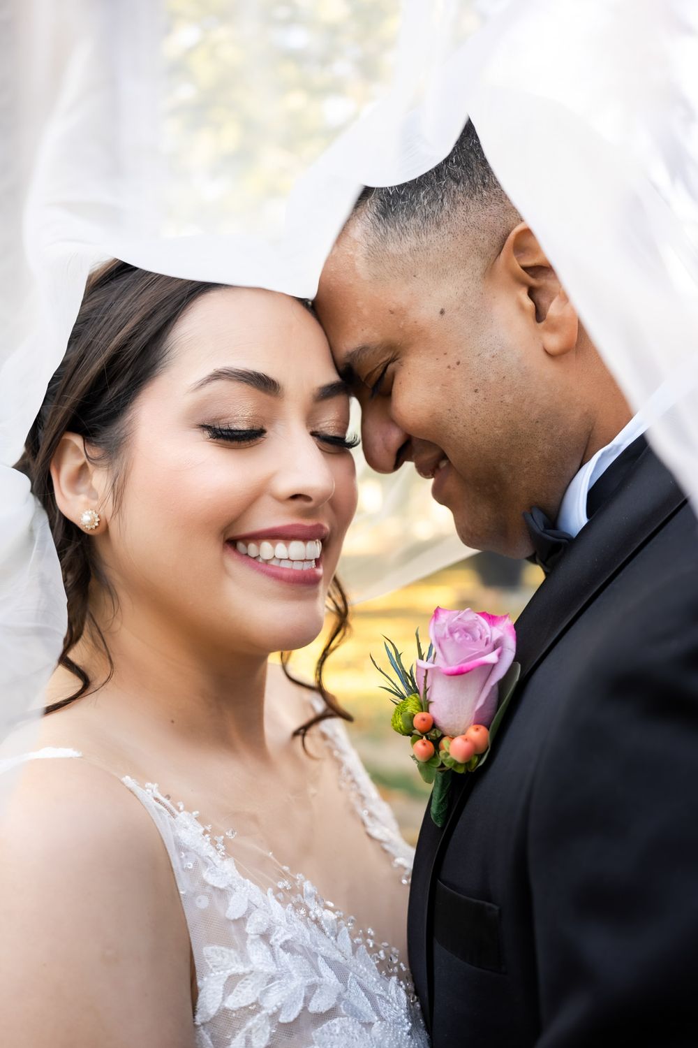 Bride smiles with closed eyes as she and groom touch foreheads tenderly under a veil.