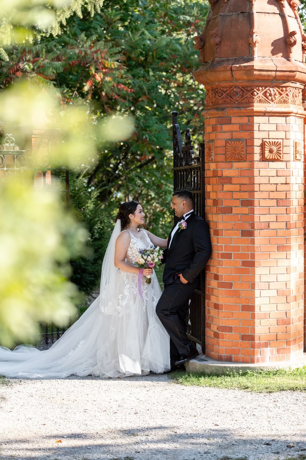 Bride and groom pose romantically beside ornate red brick tower surrounded by lush green trees.
