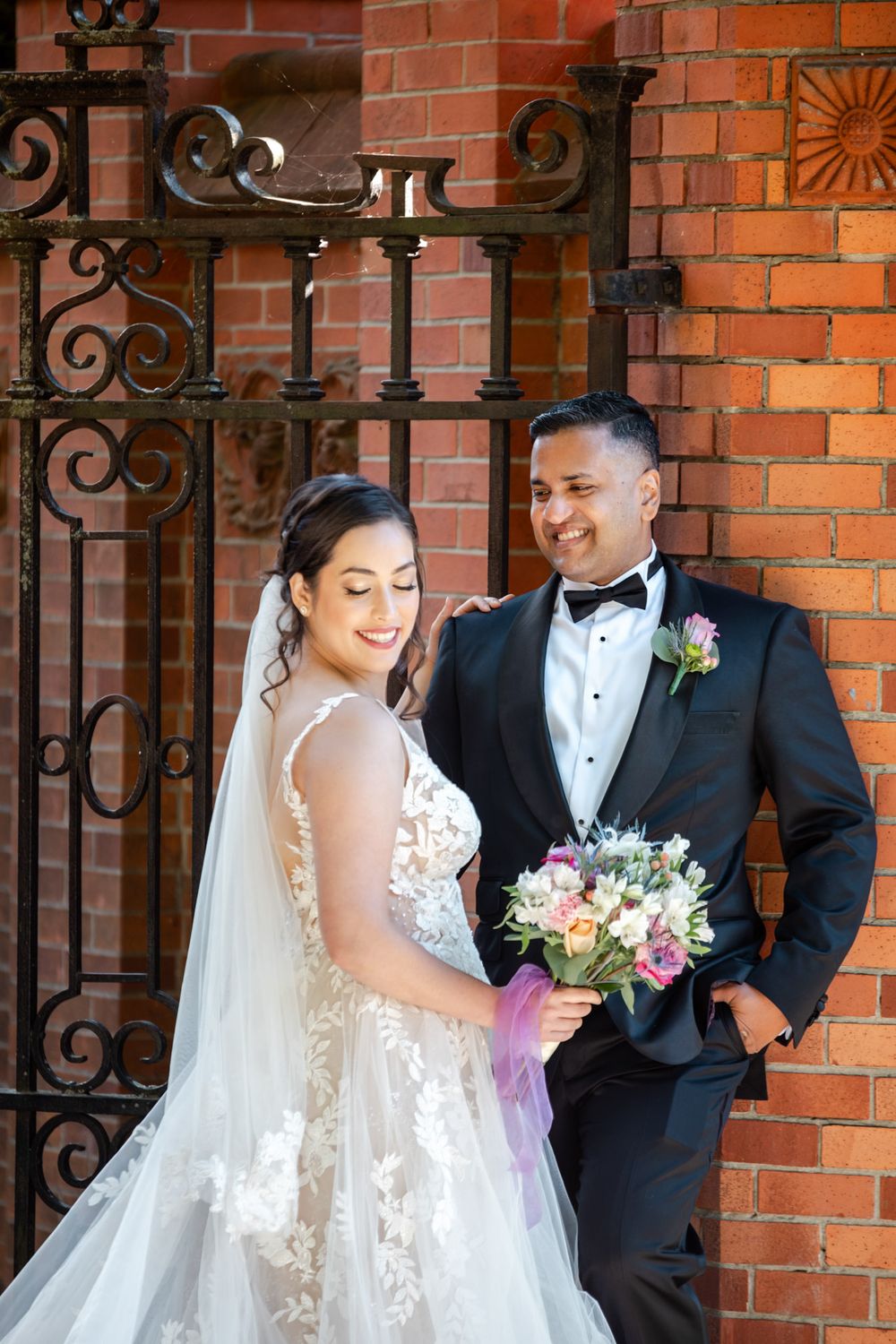 Bride and groom smiling together by ornate iron gate and brick wall, bride holding colorful bouquet in lace wedding gown.