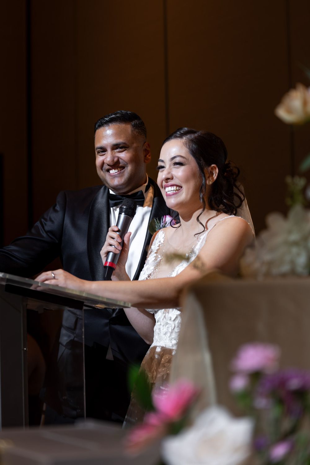 Bride and groom smile joyfully while speaking into a microphone at their wedding reception.