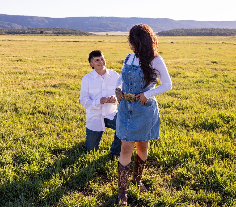Man kneeling in green field proposing to woman in denim dress and cowboy boots at golden hour.