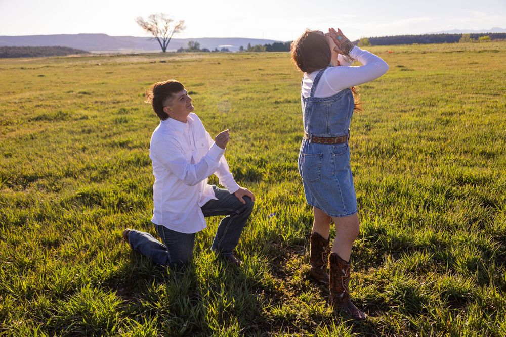 Man kneeling in green field proposing to surprised woman in denim overalls and cowboy boots at sunset.