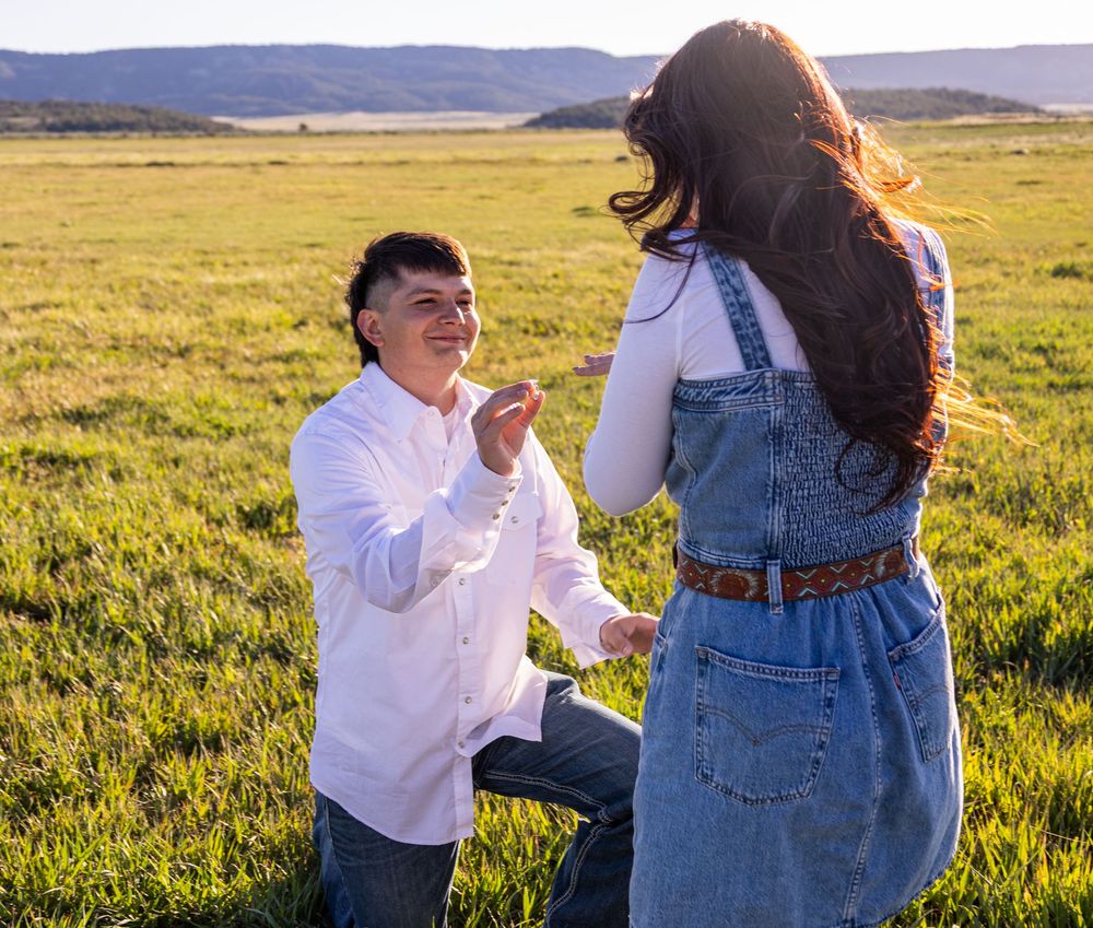 Smiling man on one knee proposing to woman in denim overalls in a sunlit green meadow.