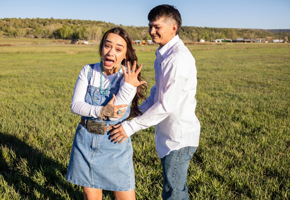 A joyful couple celebrates an engagement in a green field, woman showing off her ring while man smiles behind her.