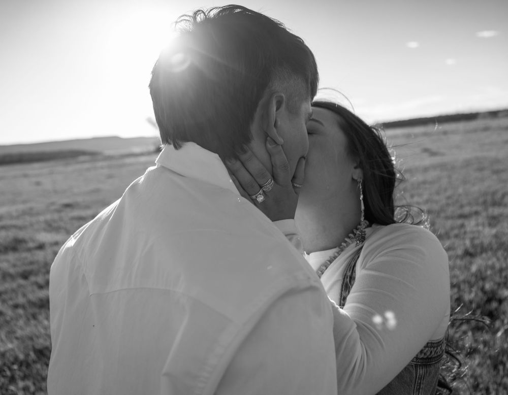 Black and white photo of a couple sharing a romantic kiss in an open field with sunlight glowing behind them.