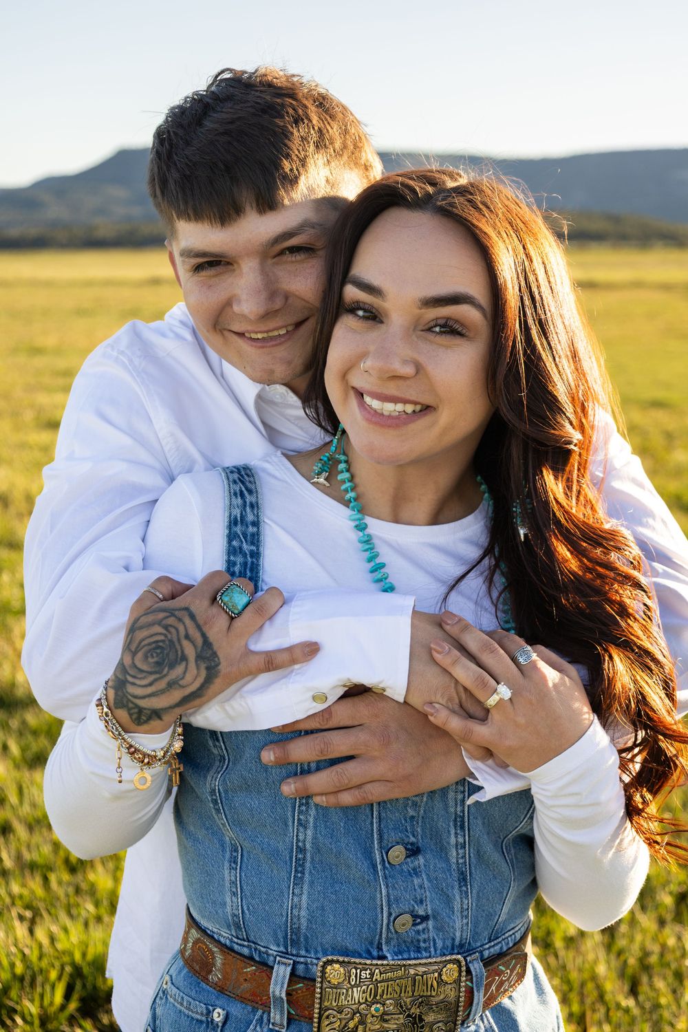 Couple embracing in a sunny green field, woman in denim overalls with turquoise necklace, man with tattoos hugging her from behind.