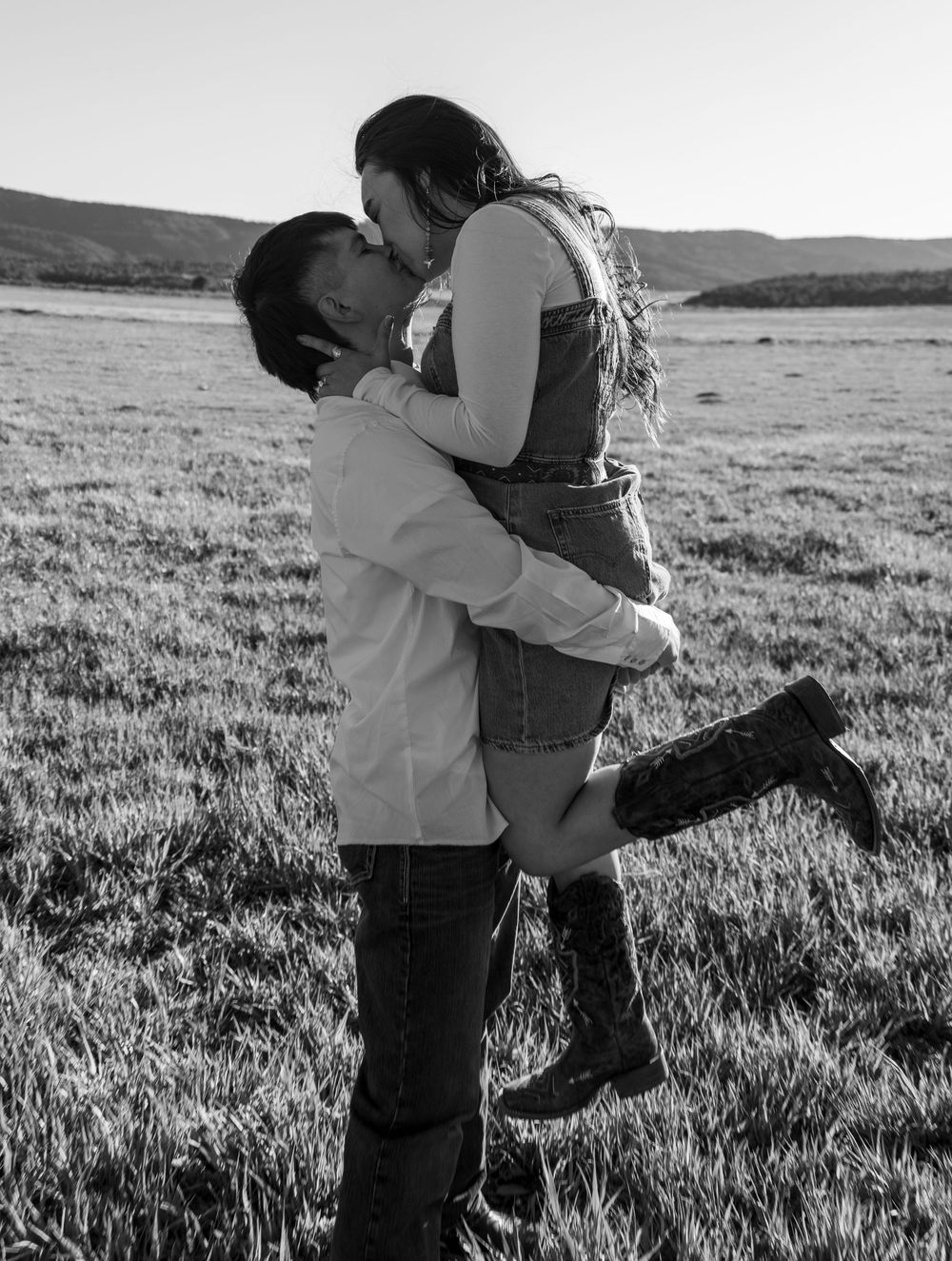 Black and white photo of a couple kissing in a field, man lifting woman wearing cowboy boots with mountains in background.