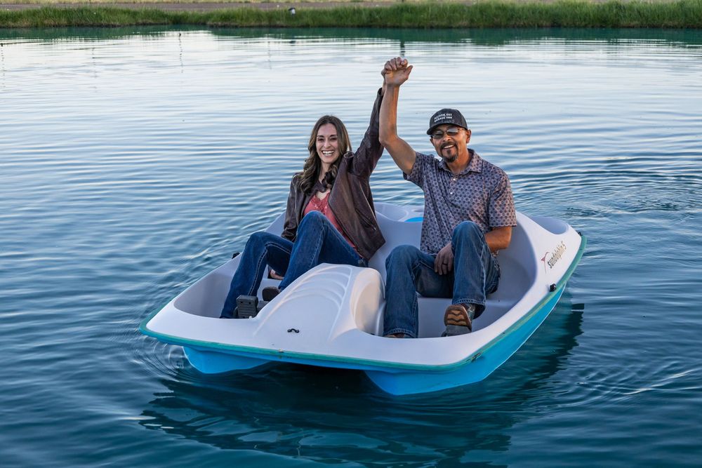 Two people smiling and celebrating on a white and teal pedal boat on a calm blue lake.
