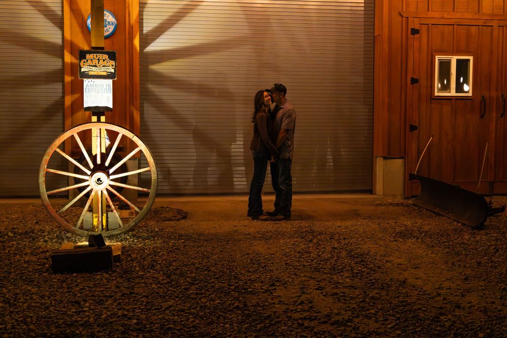 Couple sharing a romantic kiss at night beside a rustic wagon wheel outside a wooden barn.