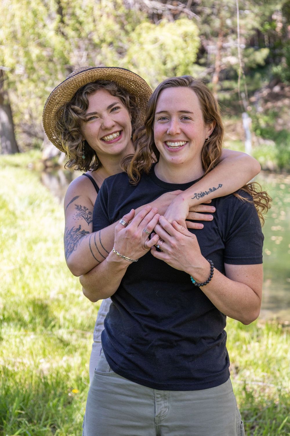 Two smiling women embrace outdoors in a sunny green park, one wearing a straw hat and both in navy shirts.