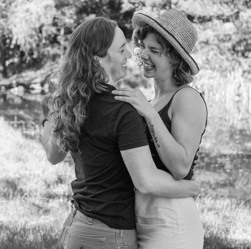 Two women laughing and embracing outdoors in a black and white photo near trees and water.