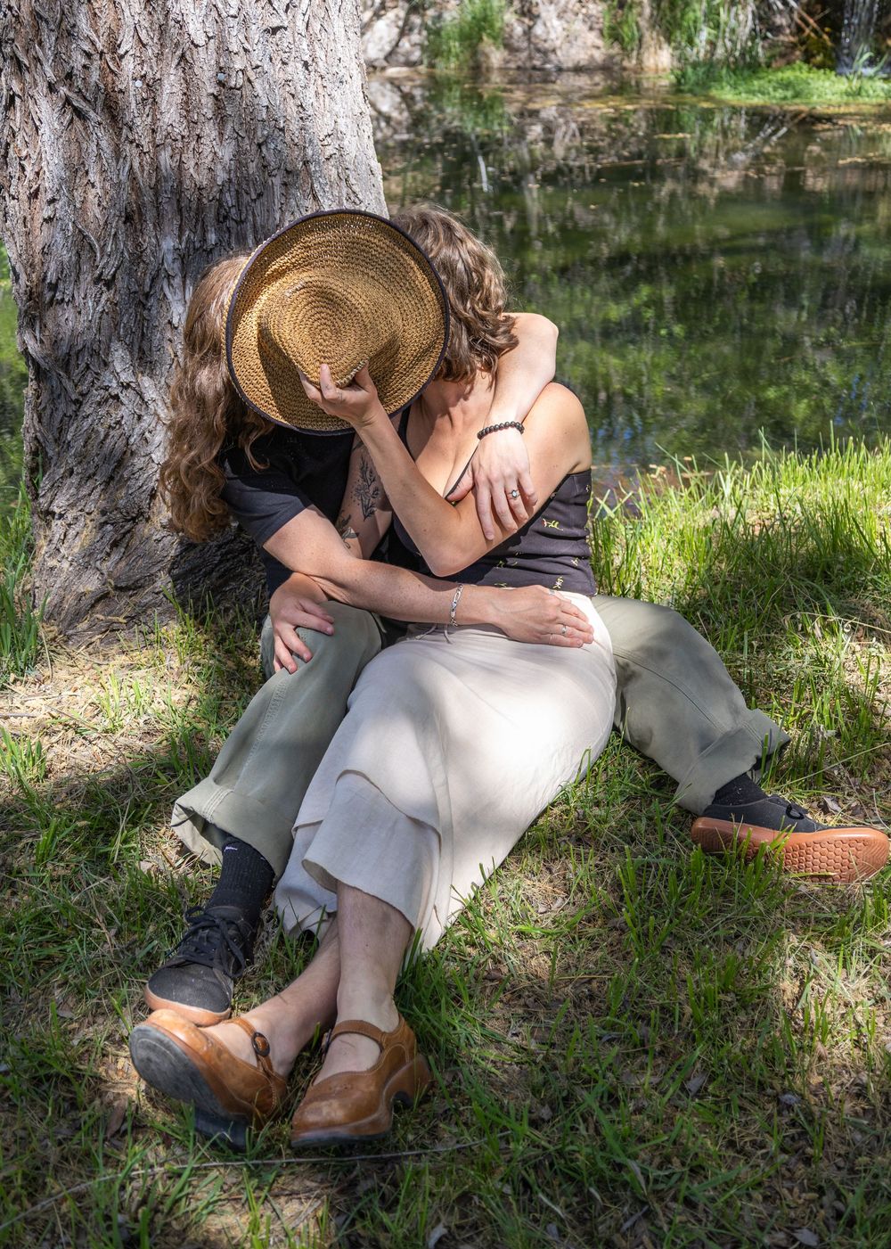 Couple embracing romantically under a tree by a pond, woman in straw hat and cream skirt, man in olive pants.