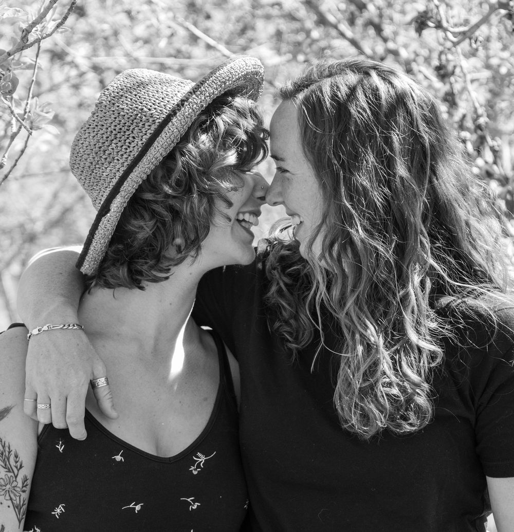 Two women smiling and laughing together outdoors, one wearing a straw hat, in a black and white photo.
