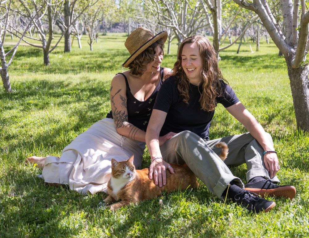 Two women sit together on green grass with an orange cat, relaxing under trees in a sunny orchard.