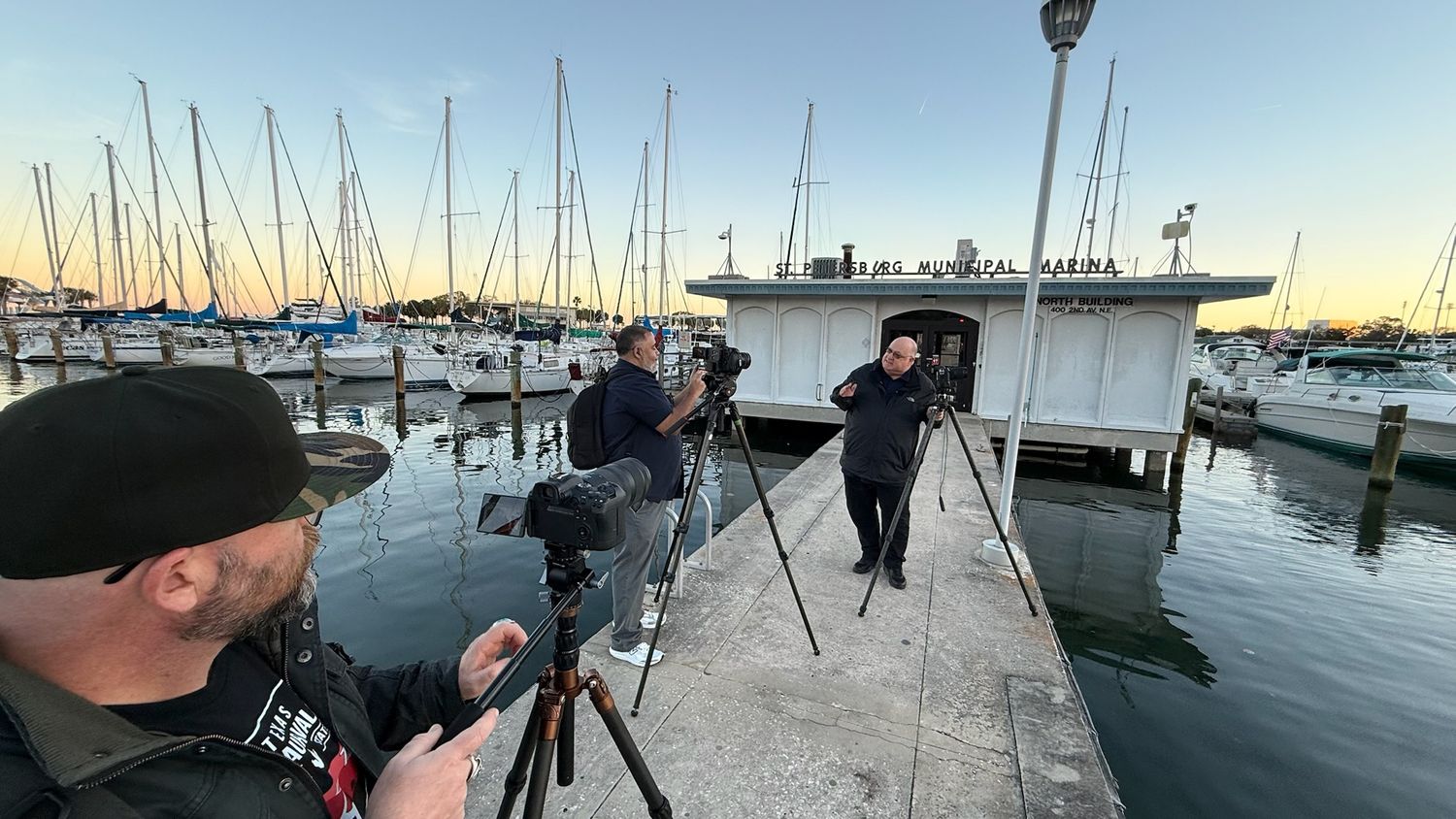 Photographers with tripods shoot golden hour at a marina with sailboats docked at sunset.