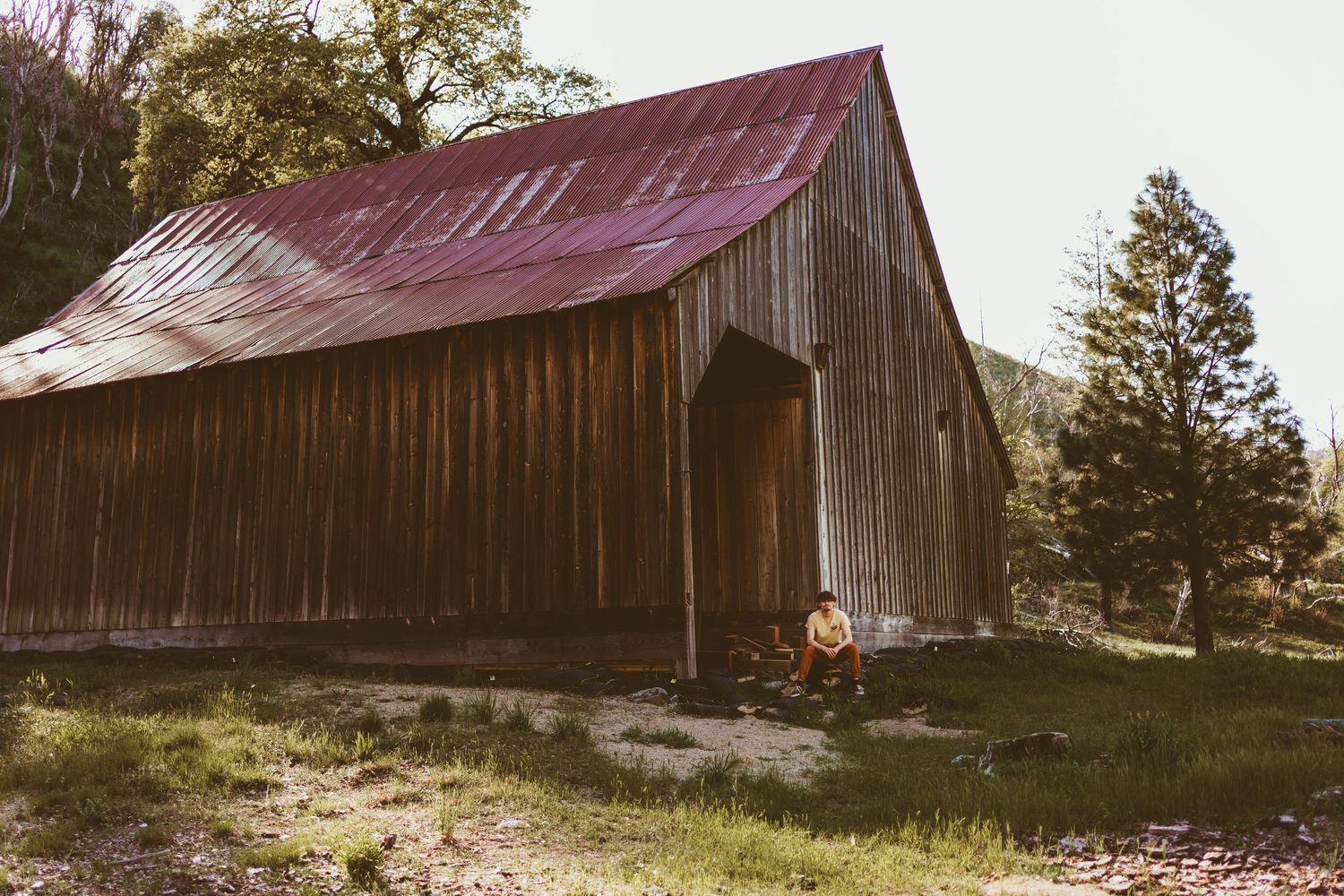 Person sitting beside a rustic wooden barn with a red metal roof surrounded by trees and green grass.