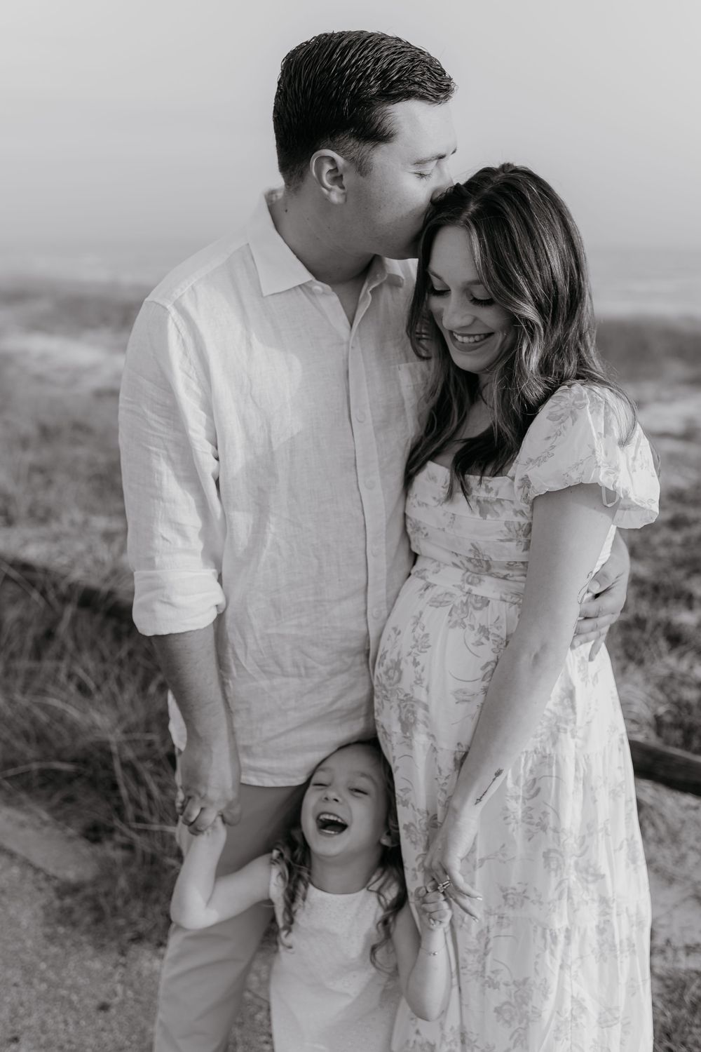 Black and white maternity photo of a couple with young daughter laughing on a beach path, woman in floral dress.