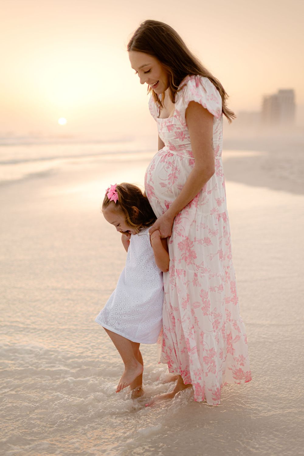 Pregnant woman in floral dress with toddler girl on beach at sunset, waves washing over their feet.