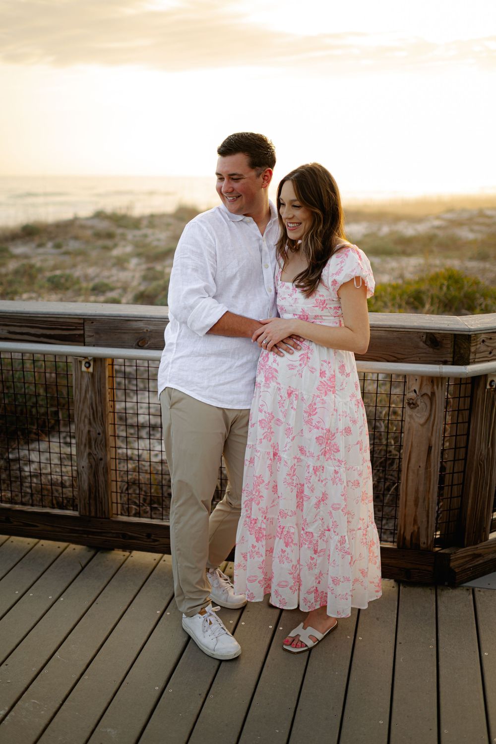Couple embracing on a wooden beach boardwalk at sunset, man in white shirt and woman in pink floral dress.